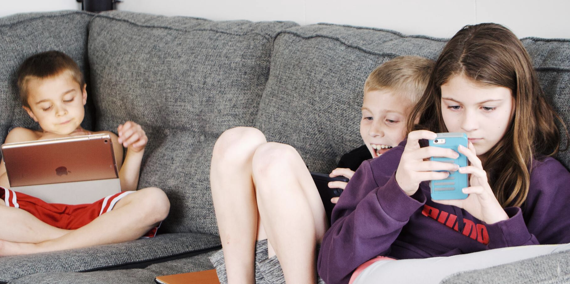 Kinder Medienkompetenz Unterschiedlich alte GEschwister sitzen auf Sofa, jedes Kind mit einem eigenen digital device.