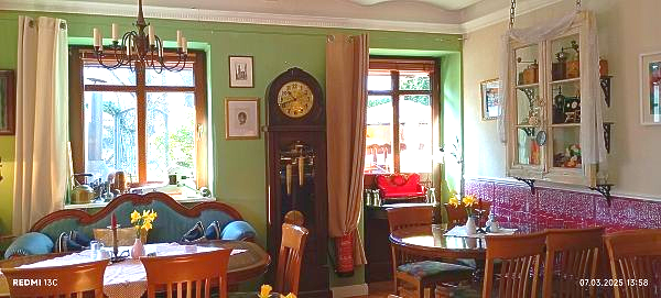 Das Café Törtchen von Innen, an der Wand steht die große Standuhr, daneben das blaue Biedermeiersofa auf der anderen Seite ein Tisch mit 4 Stühlen. es herrscht eine gemütliche Atmosphäre.
