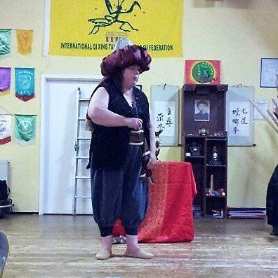 auf dem Kopf ein roter Turban, eine schwarze Weste, weiße Kurzarmbluse, blaugraue Pluderhose, Sisalpantoffeln, Auftritt in der Trainingshalle von Bulaimei Kung-Fu