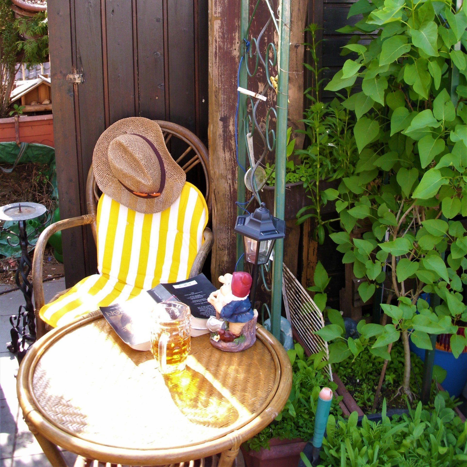 Rattan Möbel ein Sessel und ein Tisch, auf dem Tisch steht eine Lampe mit einem kleinen Zwerg, daneben steht ein Bierglas und ein Aufgeschlagenes Buch, auf dem Stuhl ist ein Cowboyartiger Hut,