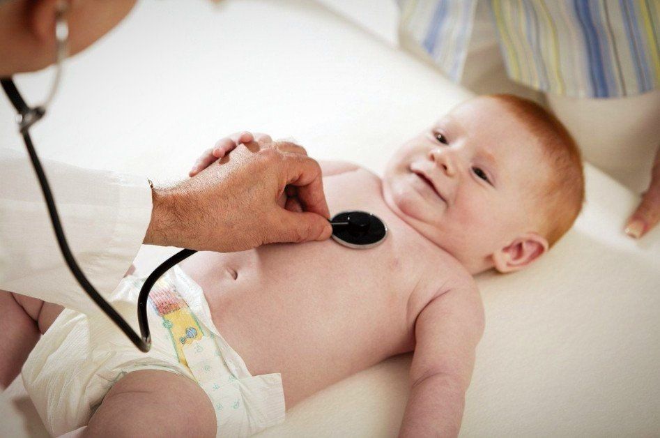 Säugling liegt in Windel auf dem Rücken auf einer Untersucherliege und wird mit einem Stethoskop an der Brust abgehört