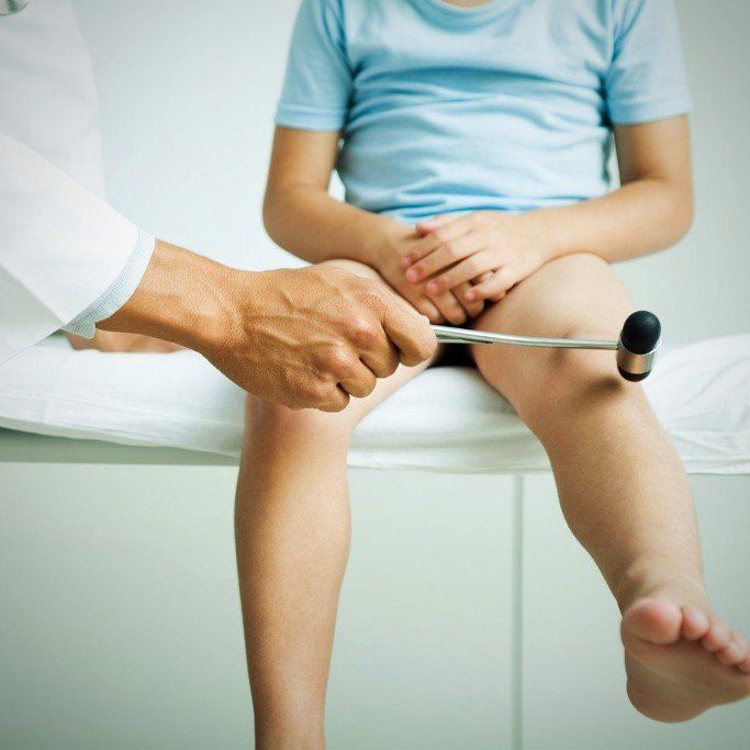 ein Kind sitzt auf einer Liege, sodass die Beine über dem Boden hängen. Die Reflexe am Knie werden mit einem Reflexhammer getestet
