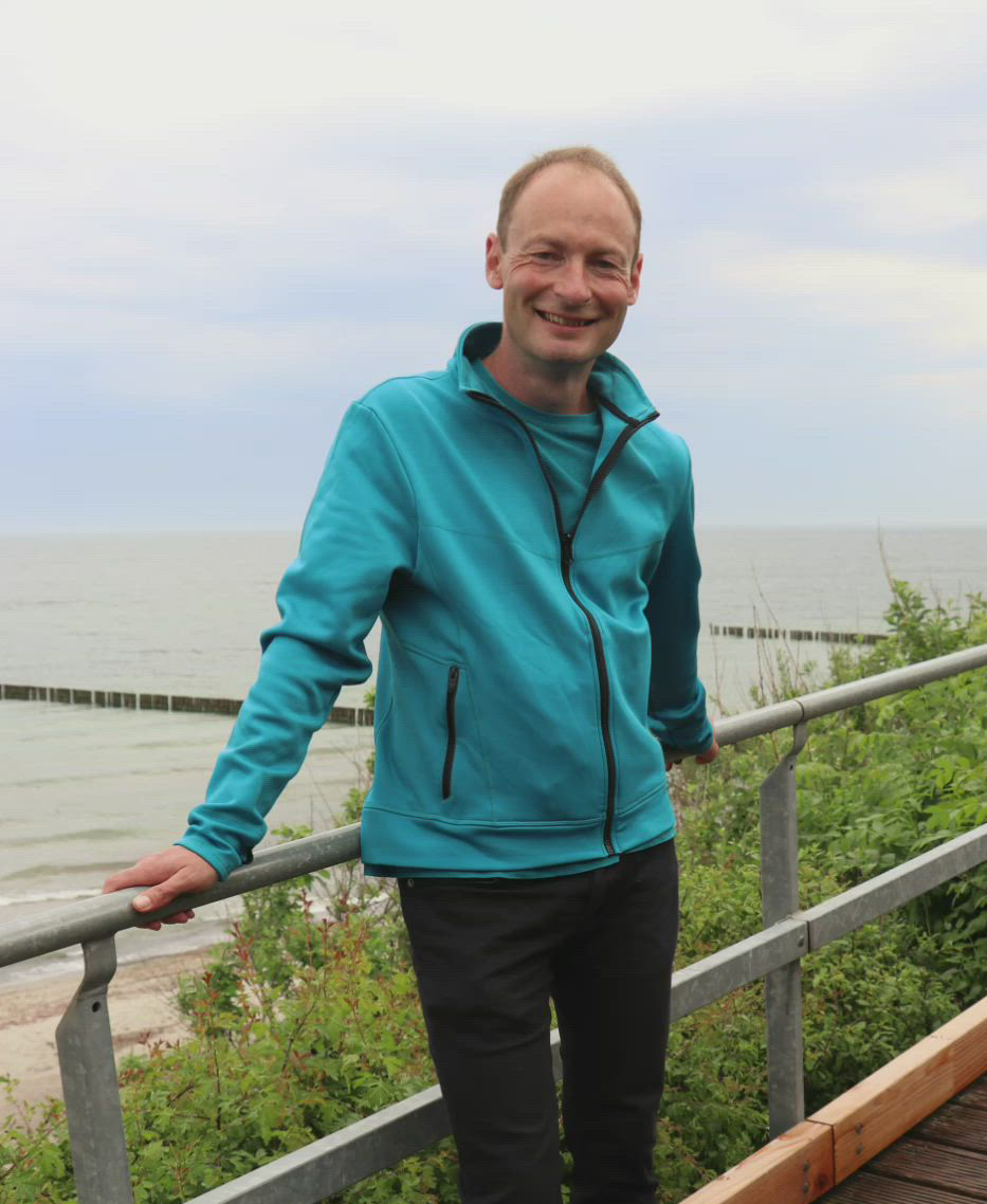 Junger Mann steht auf einer Brücke im Hintergrund die Ostsee und der Strand vom Ostseebad Nienhagen.