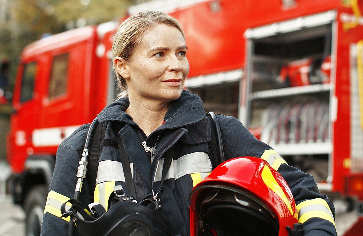 Das Bild zeigt eine Feuerwehrfrau in Arbeitskleidung mit dem Helm unter dem Arm, vor einem Einsatzfahrzeug stehend.