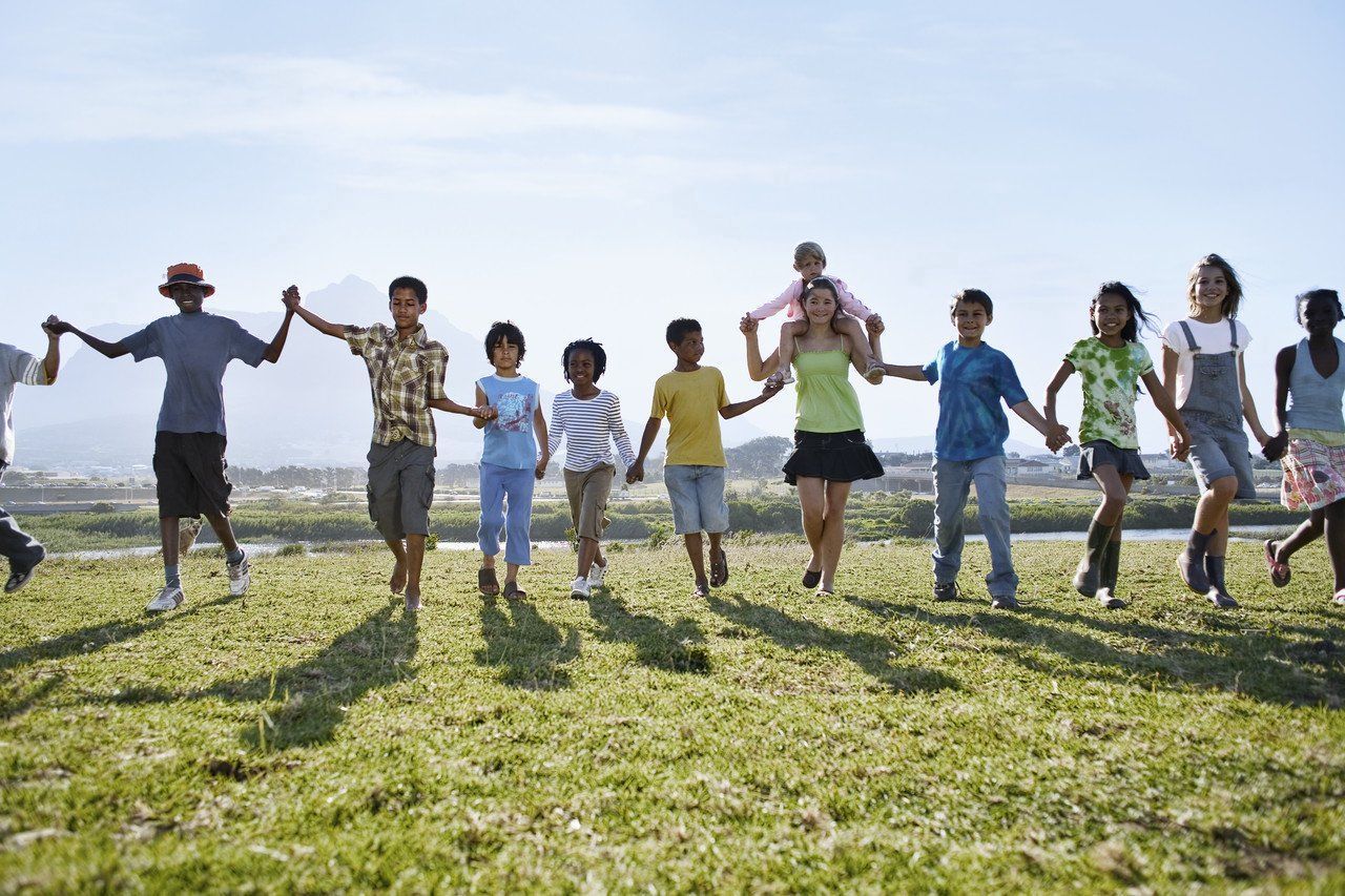 Mehrere Kinder und 1 Frau fassen sich an Händen. Kleinkind sitzt auf den Schultern der Frau. Sie gehen vorwärts auf Wiese