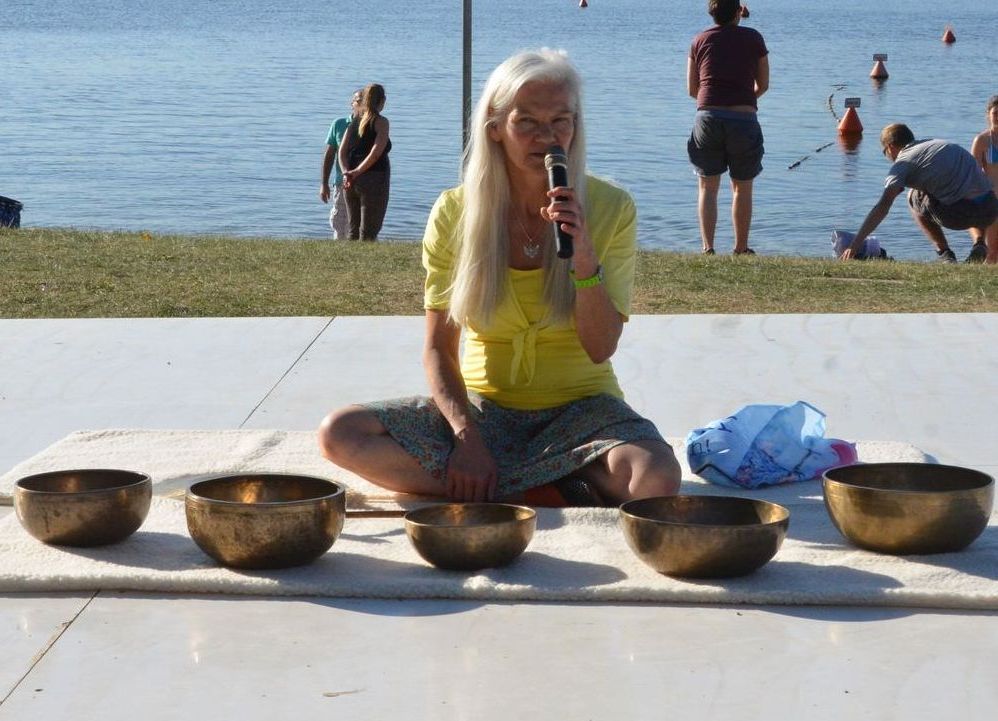 Heidrun Kirsch sitzt mit Mikro auf einer Yogamatte. Vor ihr stehen Klangschalen.