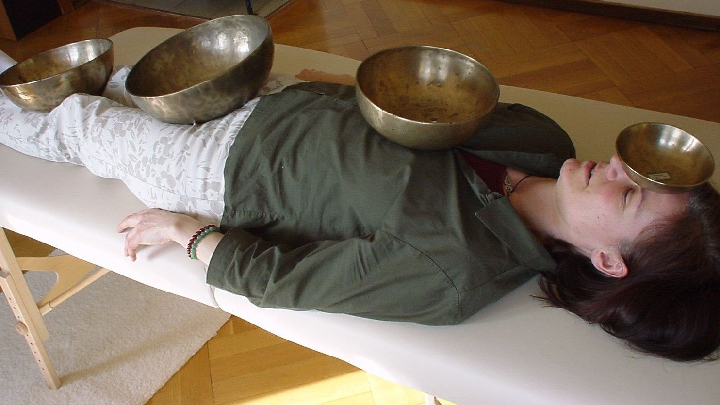 1 Frau liegt auf einer Massageliege. Klangschalen liegen verteilt auf ihrem Körper. Die frau sieht entspannt aus.