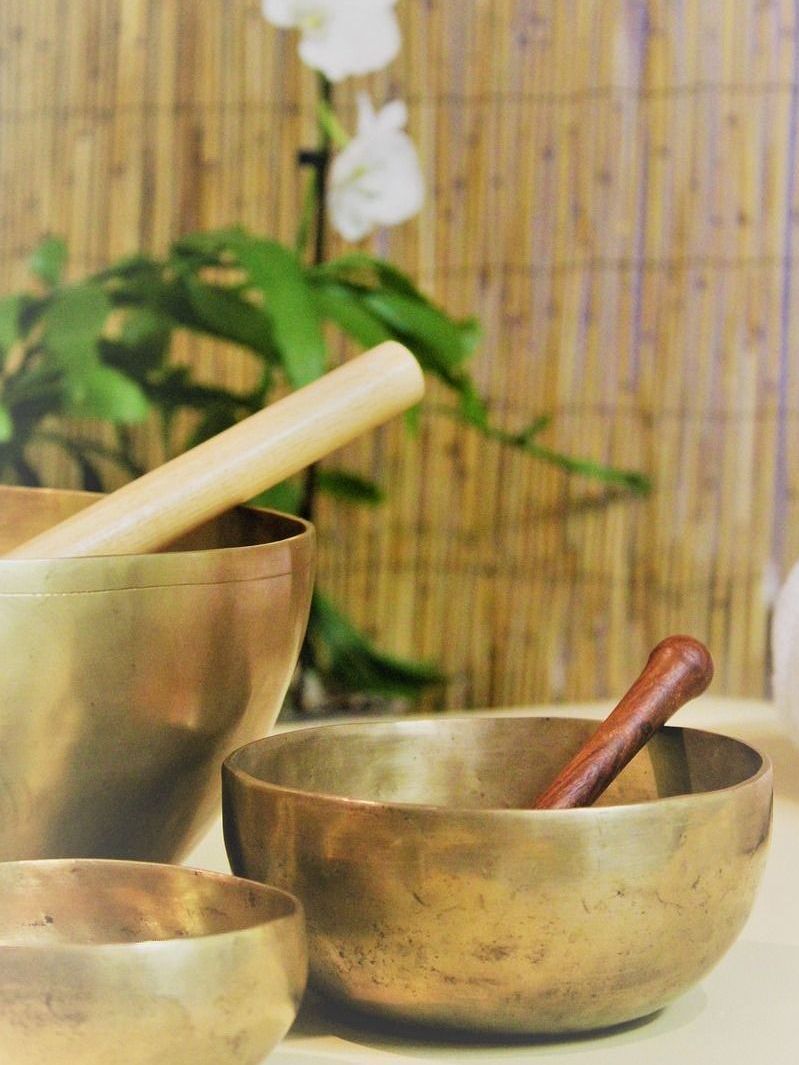 2 Klangschalen. In beiden Klangschalen ist ein Holzklöppel. Im Hintergrund ein Kunstbambus und rechts ein Bambus-Paravent.