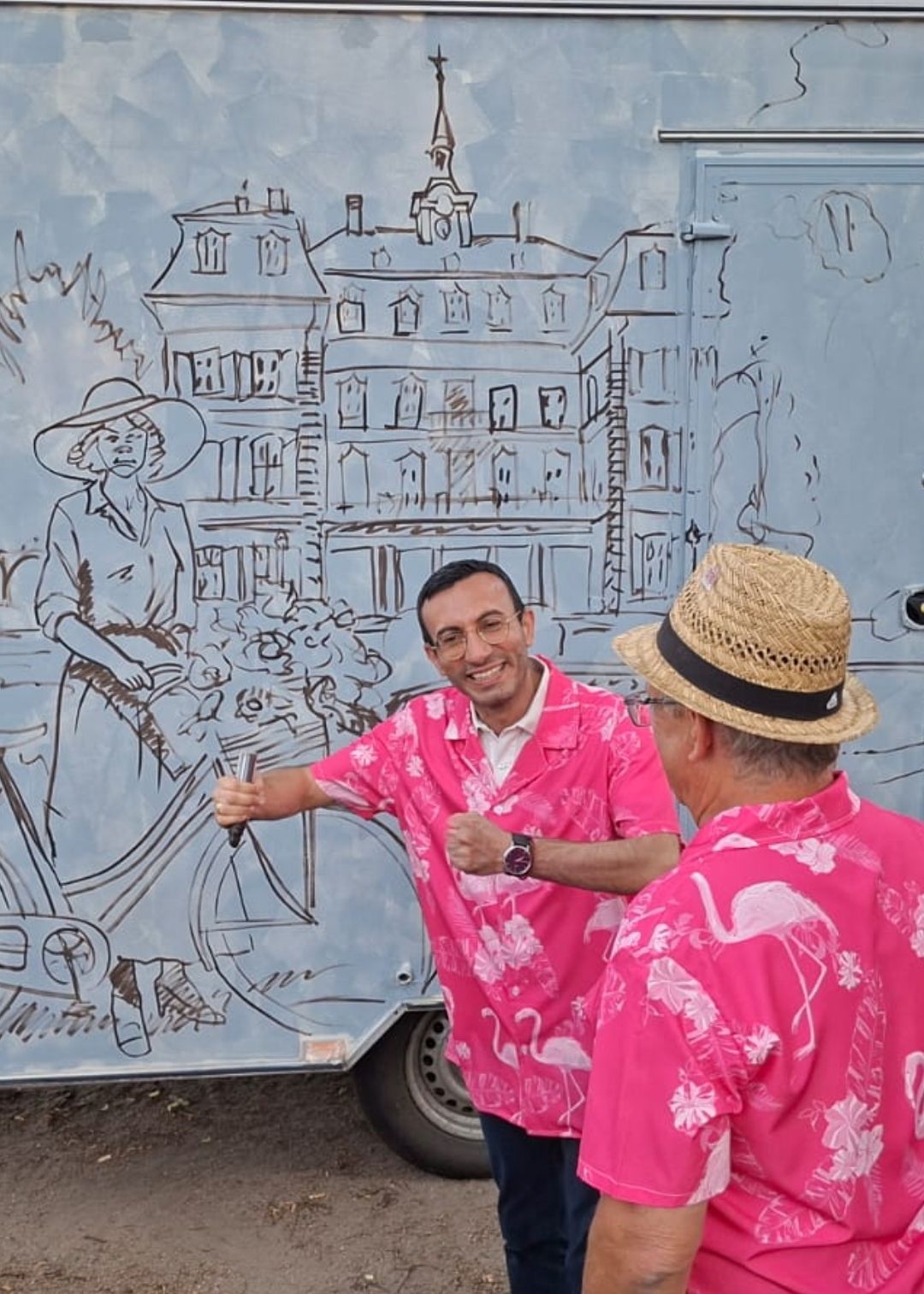 Ein Porträt von Oberbürgermeister Mike Josef (Mitte, rosa Flamingo-Hemd, Brille), der in die Kamera lächelt und den Daumen nach oben zeigt. Er steht vor dem Livepainting-Kunstwerk. Die Person rechts (im Strohhut) Claus Cromm