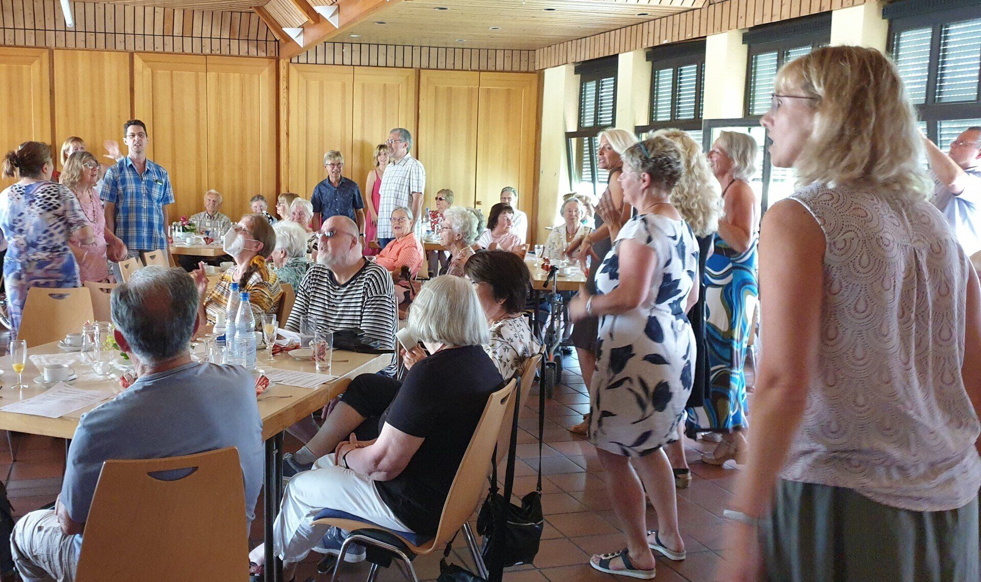 Menschen sitzen an Tischen. Dazwischen stehen Choristen und singen.
