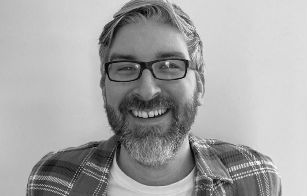Black and white image of man with beard and glasses with flannel shirt in front of blank wall