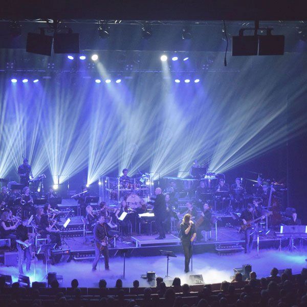 Konzert Bremen Die Band und das Orchester von Philharmonic Rock spielen ein Konzert auf der Bühne des Metropol Theaters. Scheinwerfer leuchten nach vorne.