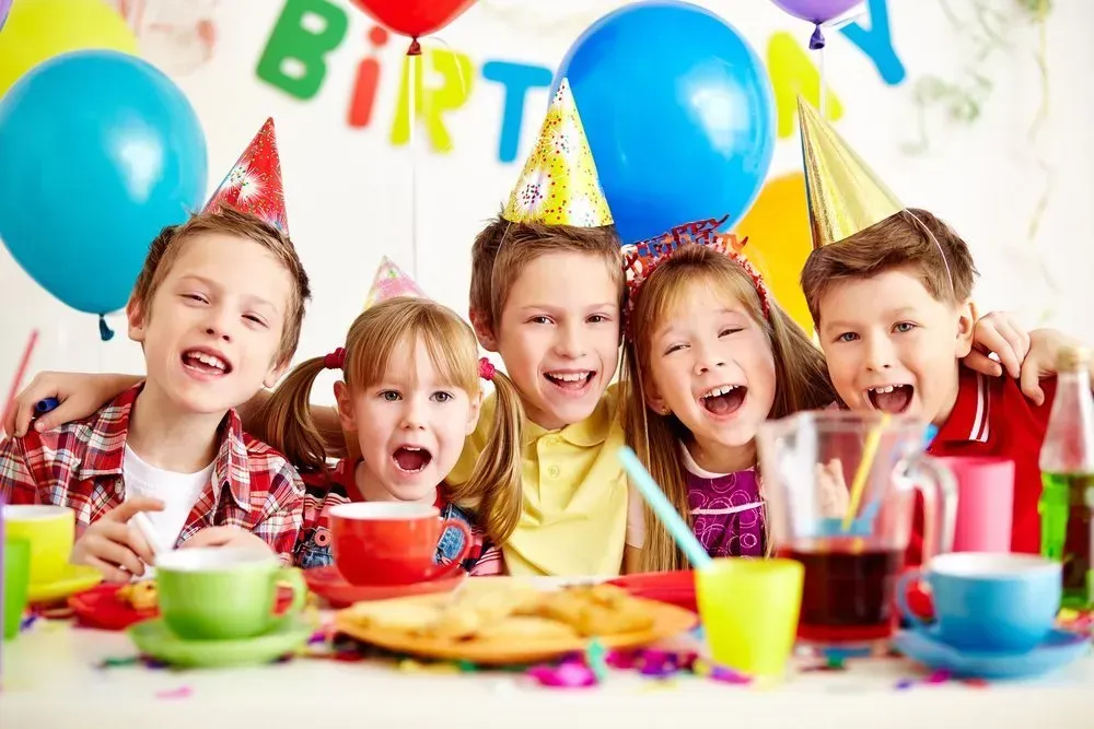Bild Kindergeburtstag. Kinder feiern am Geburtstagstisch mit Ballons, Luftschlagen und Konfetti im Hintergrund