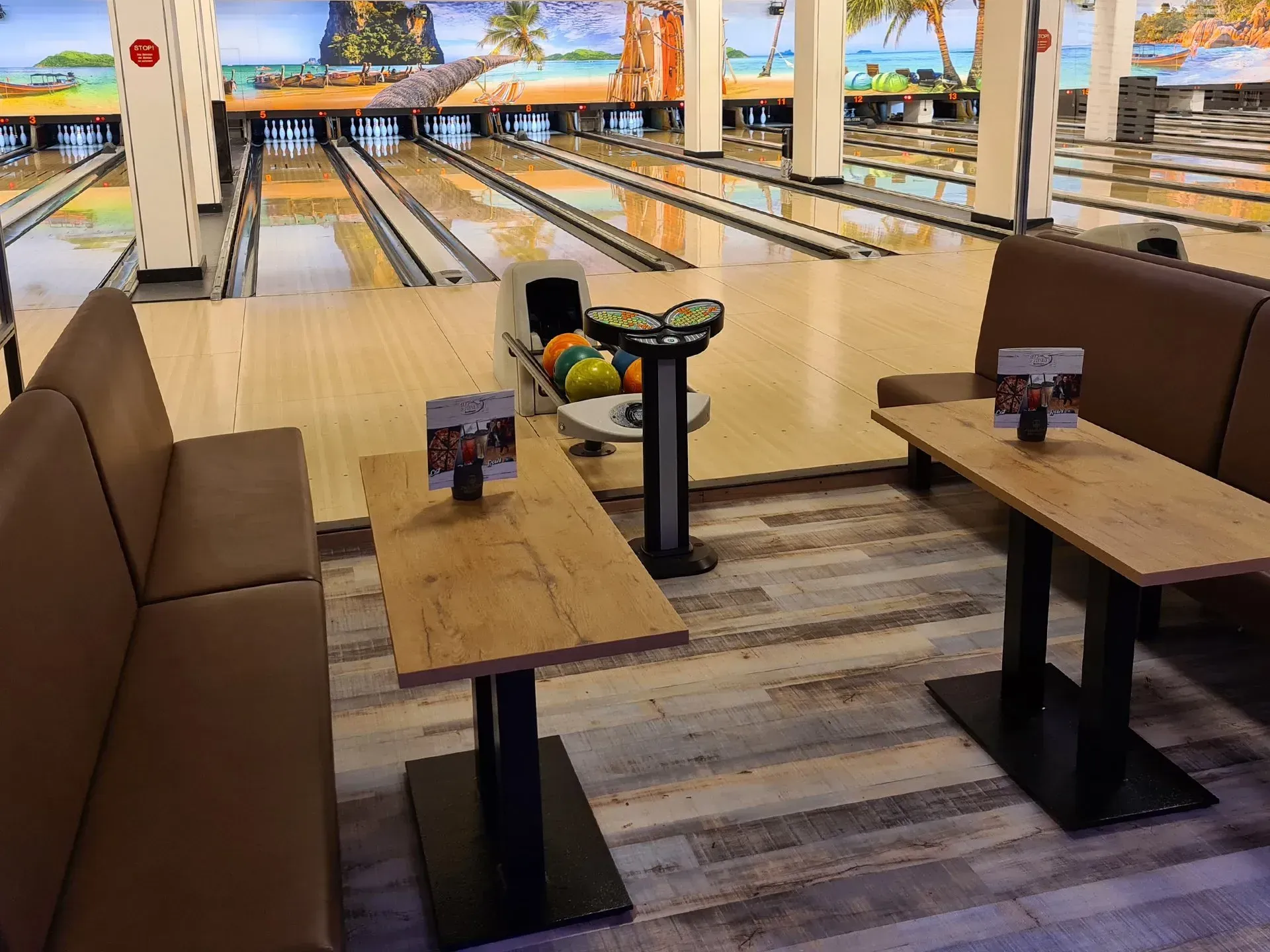Bild unserer Bowlingbahnen mit Strandbild über den Pins. Im Vordergrund die Sitzbänke in braunem Leder und die Tische auf denen unsere Speisekarten stehen.