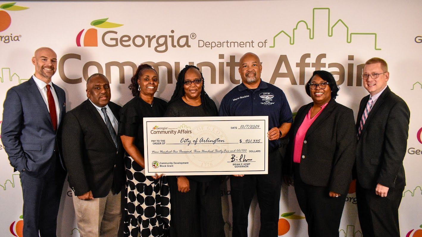 Explore our Grant Writing & Administration services Group holding a large grant check in front of Georgia Department of Community Affairs backdrop.