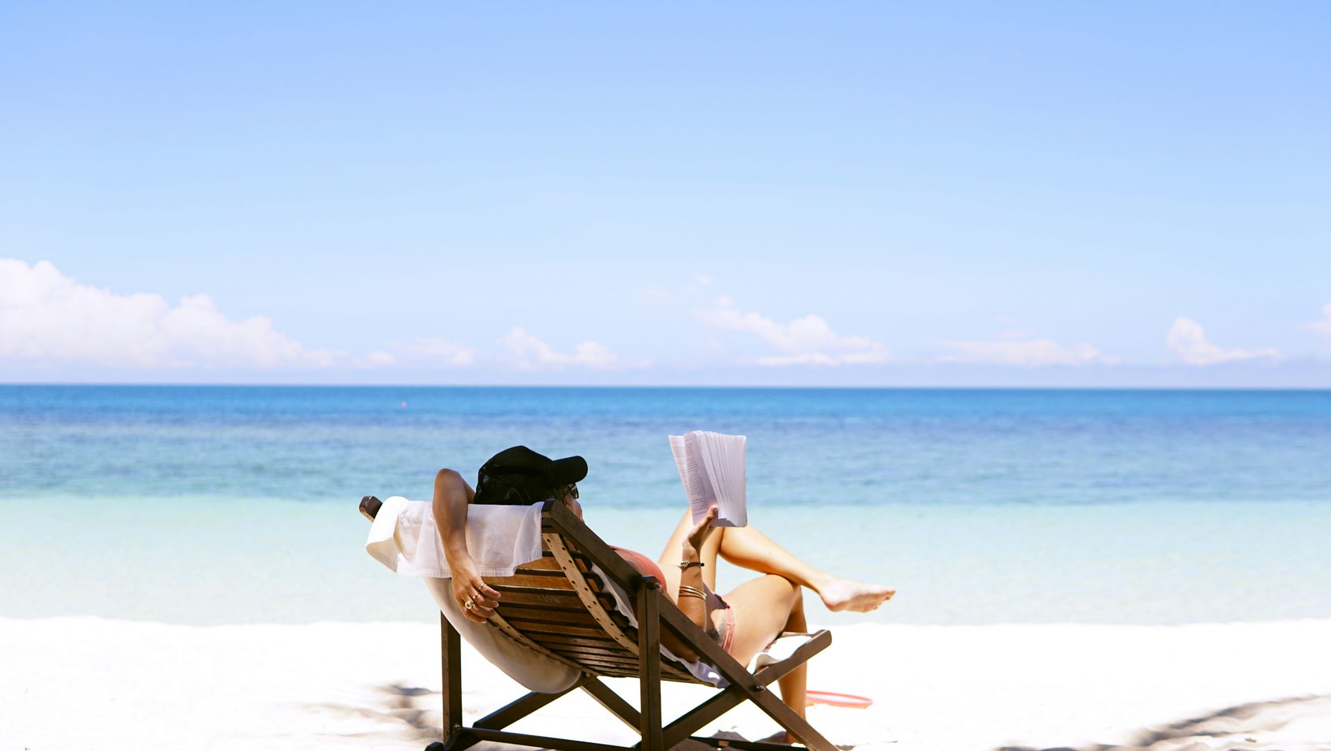 Eine Frau liest ein Buch am Strand und liegt in einem Liegestuhl, Ansicht von hinten auf Meer