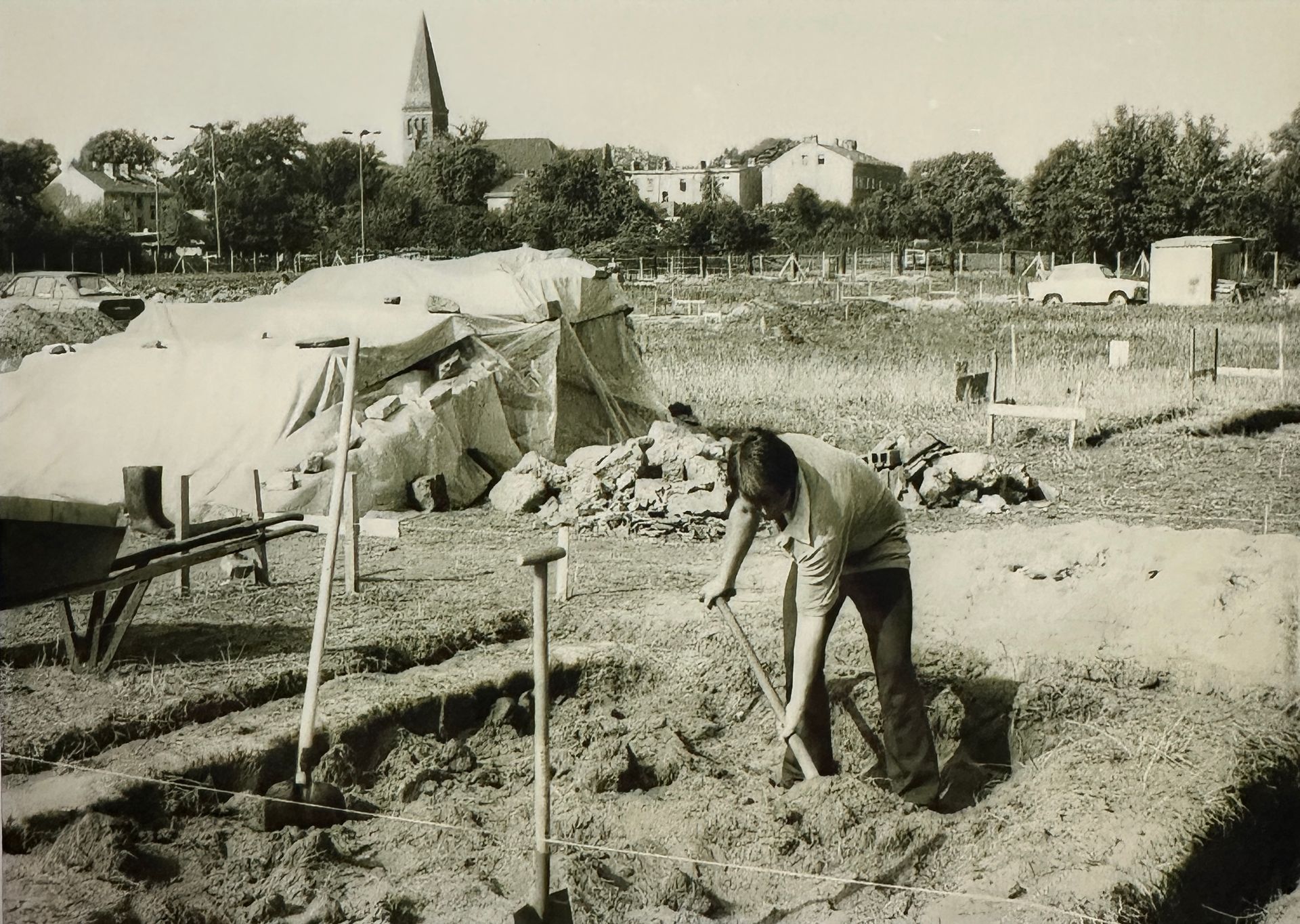 Mann steht auf KGA Gelände in einem abgegrenzten Erdstück und hebt ein Loch mit einem Spaten aus. Neben ihm stecken noch 2 weitere Spaten im Boden. Im Hintergrund liegen ein paar Steintrümmer und mit Folie abgedeckte Gegenstände (nicht definiert)