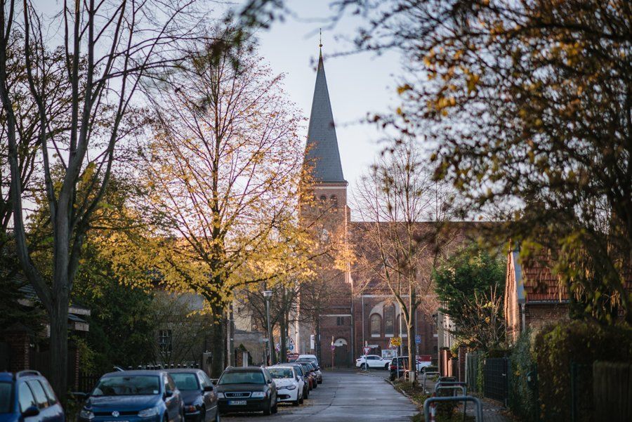 Foto der Altglienicker Kirche von weitem. Es parken ein paar Autos in der Straße vor der Kirche und Links und rechts von der Straße stehen Bäume (die Kirche am Ende, in der Mitte)
