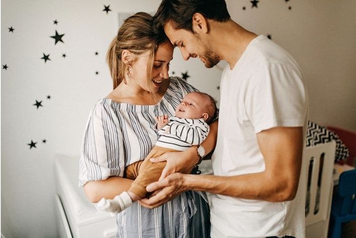 Frisch gebackene, glückliche Eltern mit ihrem Baby in den Armen, im Hintergrund sieht man das Babybettchen.