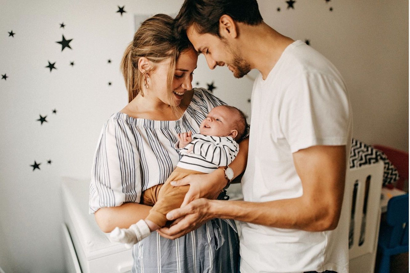 Frisch gebackene, glückliche Eltern mit ihrem Baby in den Armen, im Hintergrund sieht man das Babybettchen.
