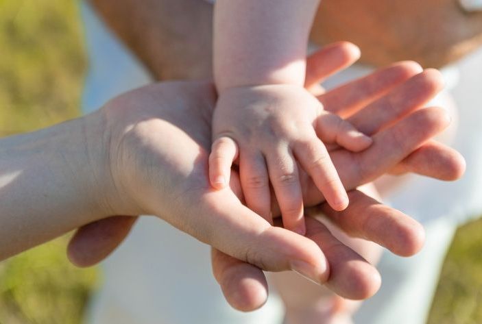 Drei ineinander liegende Hände: beide Eltern und ihr Kleinkind vereint zu einer Familie, gefeiert bei einer freien Taufe.