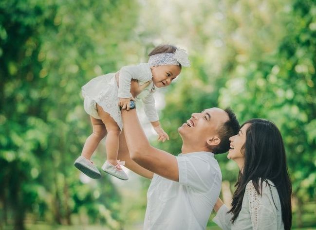 Junge Familie nach freier Taufe: Vater hält seine kleine Tochter hoch in die Luft. Vater, Mutter und Tochter strahlen.