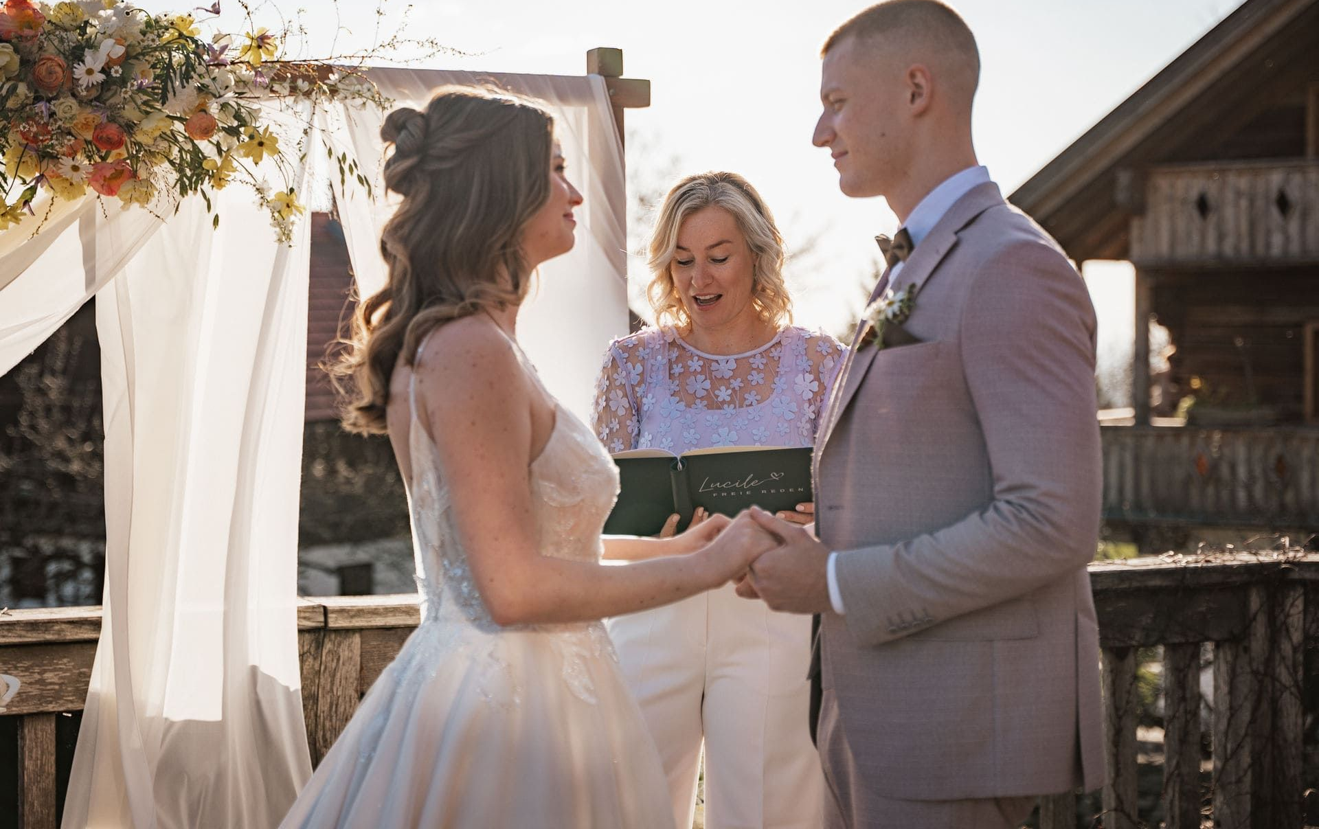 Im Vordergrund steht ein Brautpaar, das sich ansieht, im Hintergrund Lucile, die die Traurede hält.