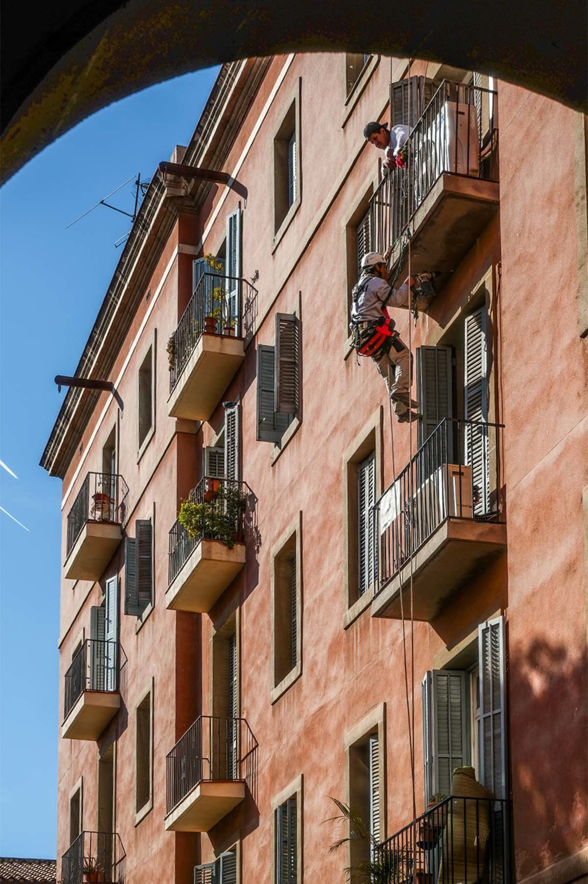 Perspectiva exterior desde el ángulo lateral a través de la arcada del edificio colindante, donde se muestra el trabajo de rehablitación de la fachada con el operario colgando para realizar el trabajo de reparación de forjado del balcón. Proporción vertical