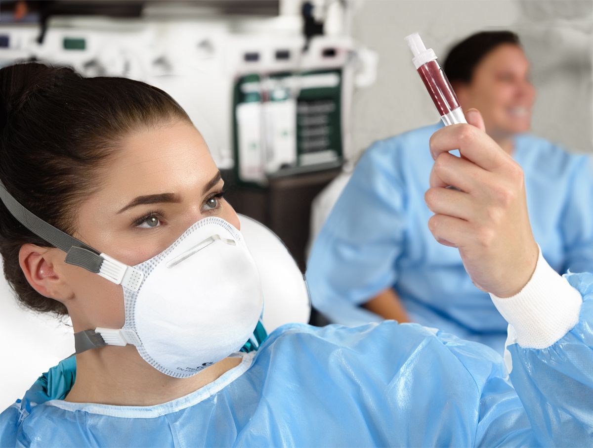 Junge Frau mit Atemschutzmaske im Labor und betrachtet eine Blutprobe gegen das Licht.