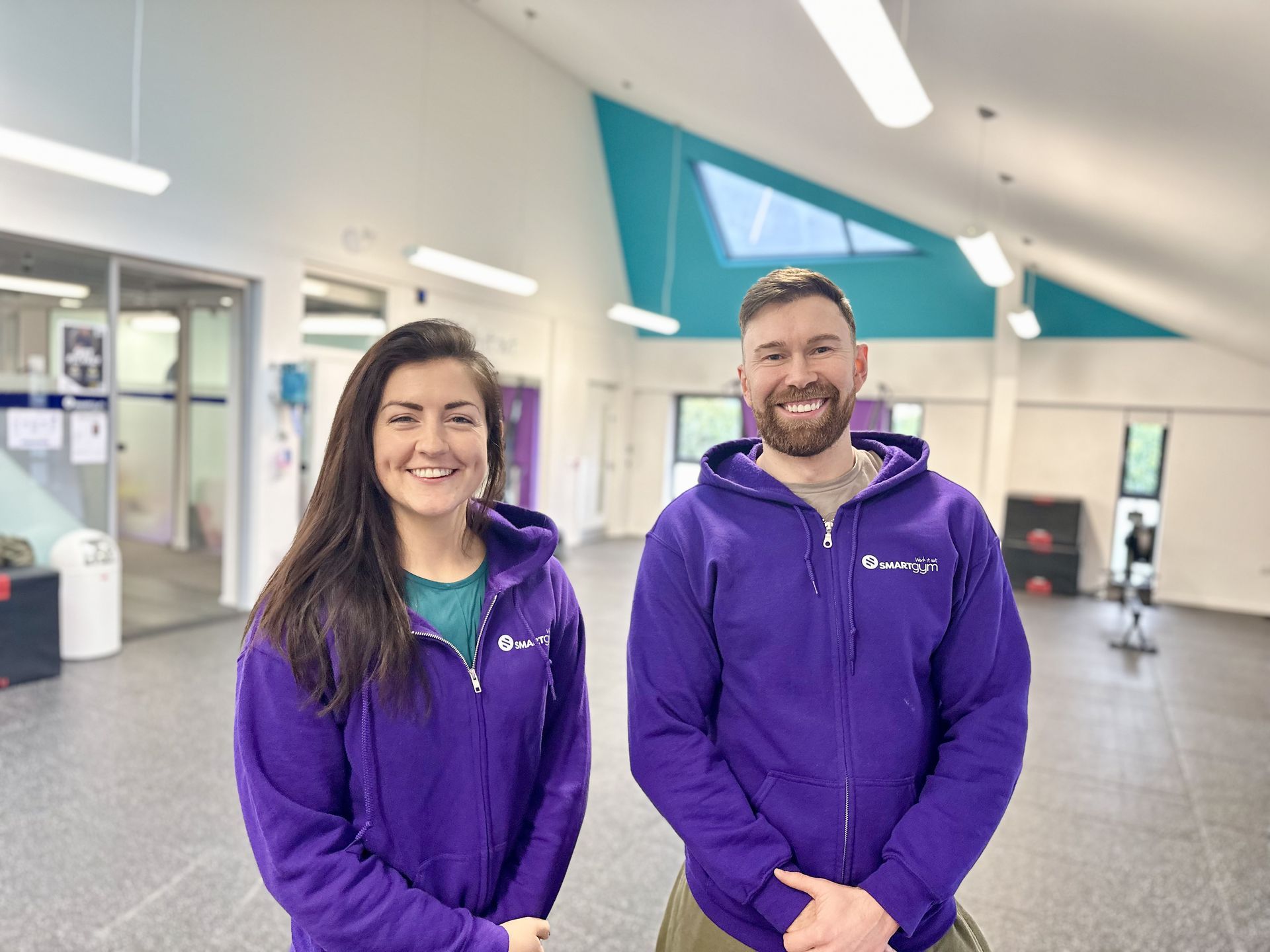 Our PT L3 tutors, Jenny and Gary. Glasgow PT Academy tutors standing side by side wearing purple Smart Gym hoodies inside SmartGym at The Wedge in Pollok