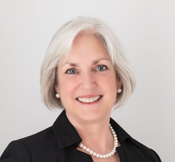 A photograph of Sara Price Celebrant, a woman with white hair wearing pearls and a black jacket, smiling
