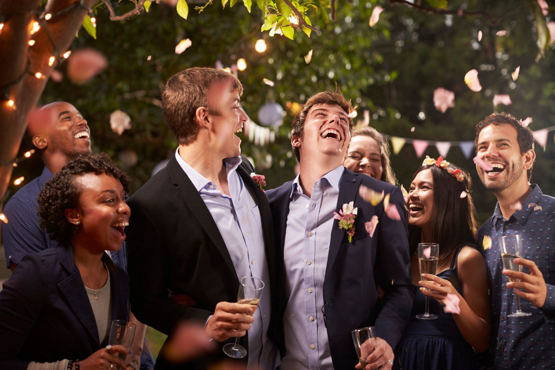 Groom and groom celebrating after their wedding ceremony laughing with and surrounded by their friends under falling rose petals.