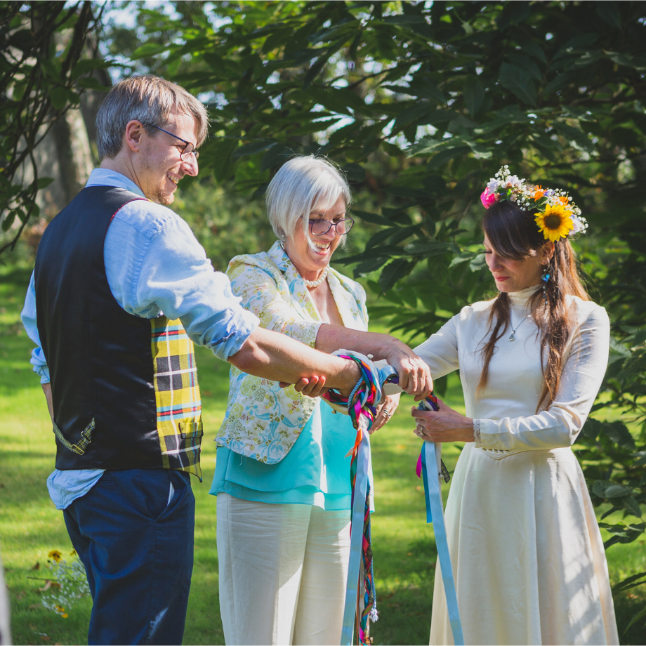 Sara Price Celebrant tying the knot around bride and groom's wrists during a hand tying ceremony beneath a tree in the garden of the wedding venue.