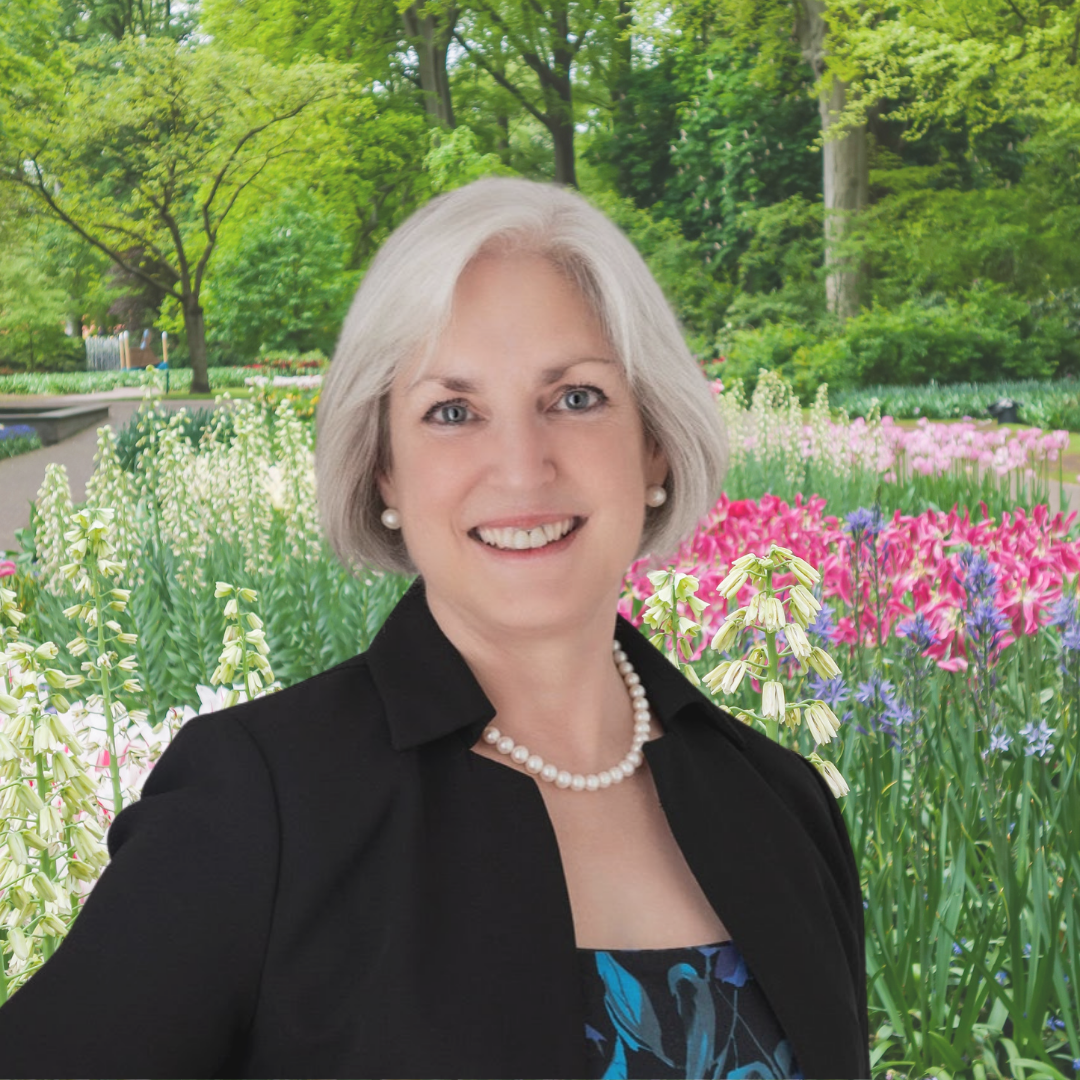 Sara Price Celebrant wearing a black jacket over a blue and black dress, with pearl necklace and earrings. She is standing in front a large bed of pink, white and blue flowers with trees in the background.