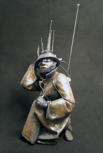 a crouching figure , swathed in protective clothes insxribed with circuit diagrams, and wearing a colander-helmet covered in antennas