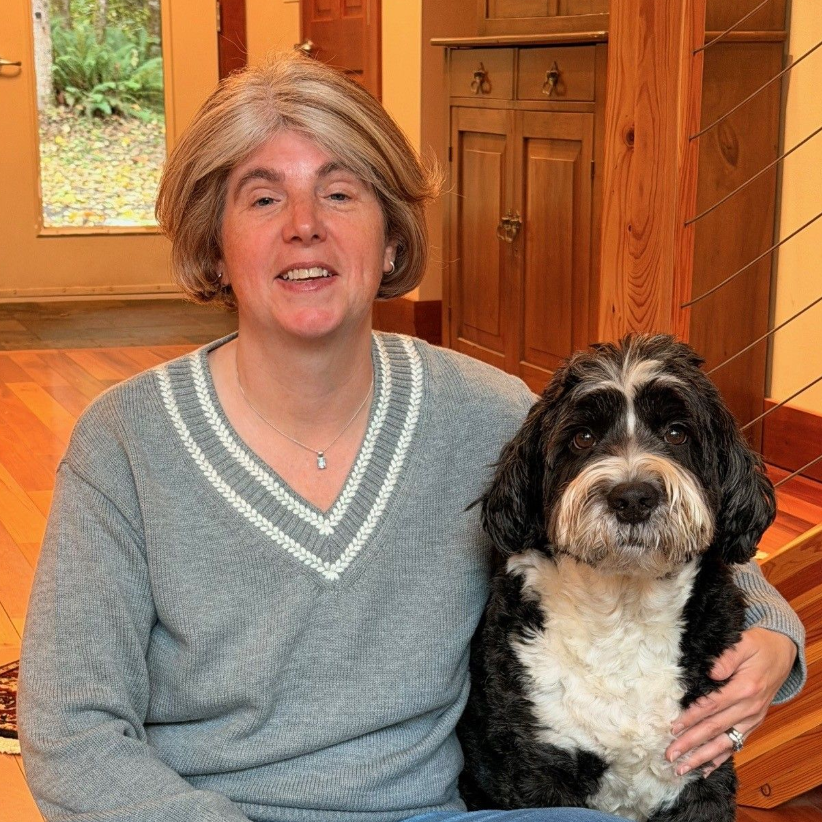 Dr. Rogers, a lady in a grey sweater sitting on the wood floor with her bernedoodle, Molly.