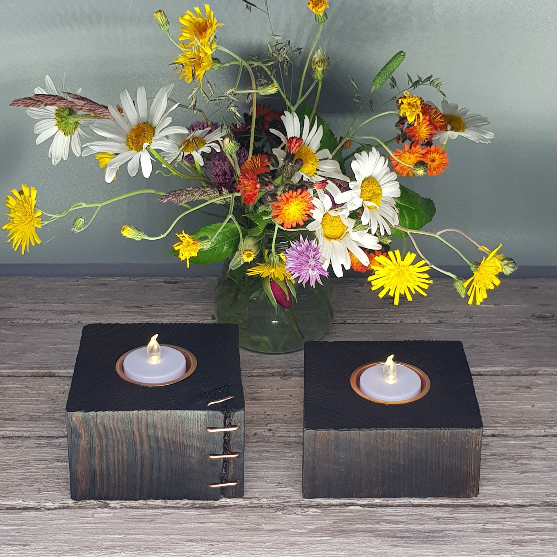 Two tealight holders made from old fence posts, positioned on a weathered wooden surface with some cut wild flowers in the background.