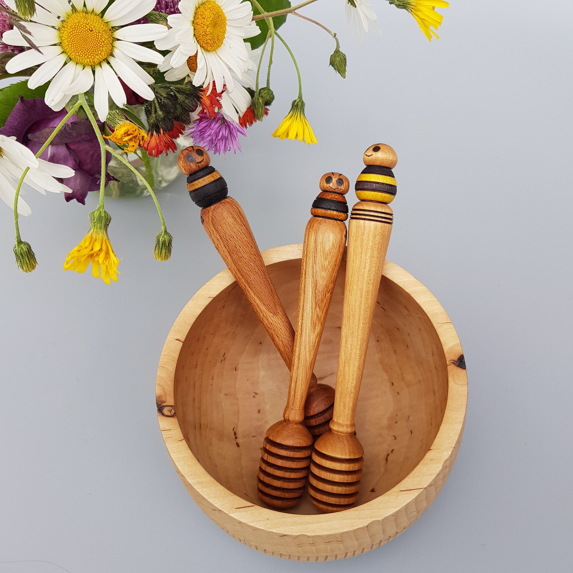 Wooden honey dippers topped with deocrated wooden bee. Placed in a woodturned bowl with plain pale grey backdrop and some wildflowers in top left of image.