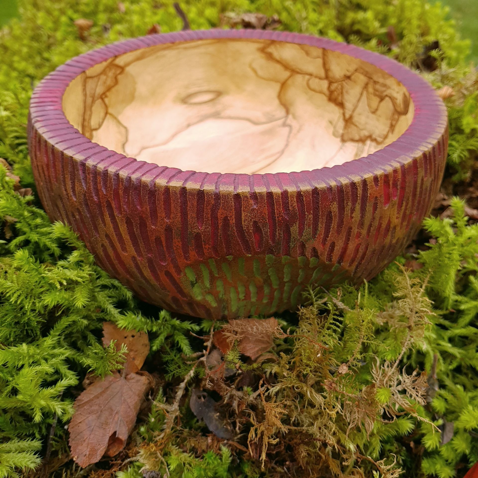 Spalted Birch Bowl Deep wooden bowl, with a textured pattern on the outside, decorated with Autumn reds. There is a depression in the shape of a leaf towards the bottom, which has been coloured green. The inside of the bowl has been left natural to show the spectacular grain pattern. It is nestled on some moss.