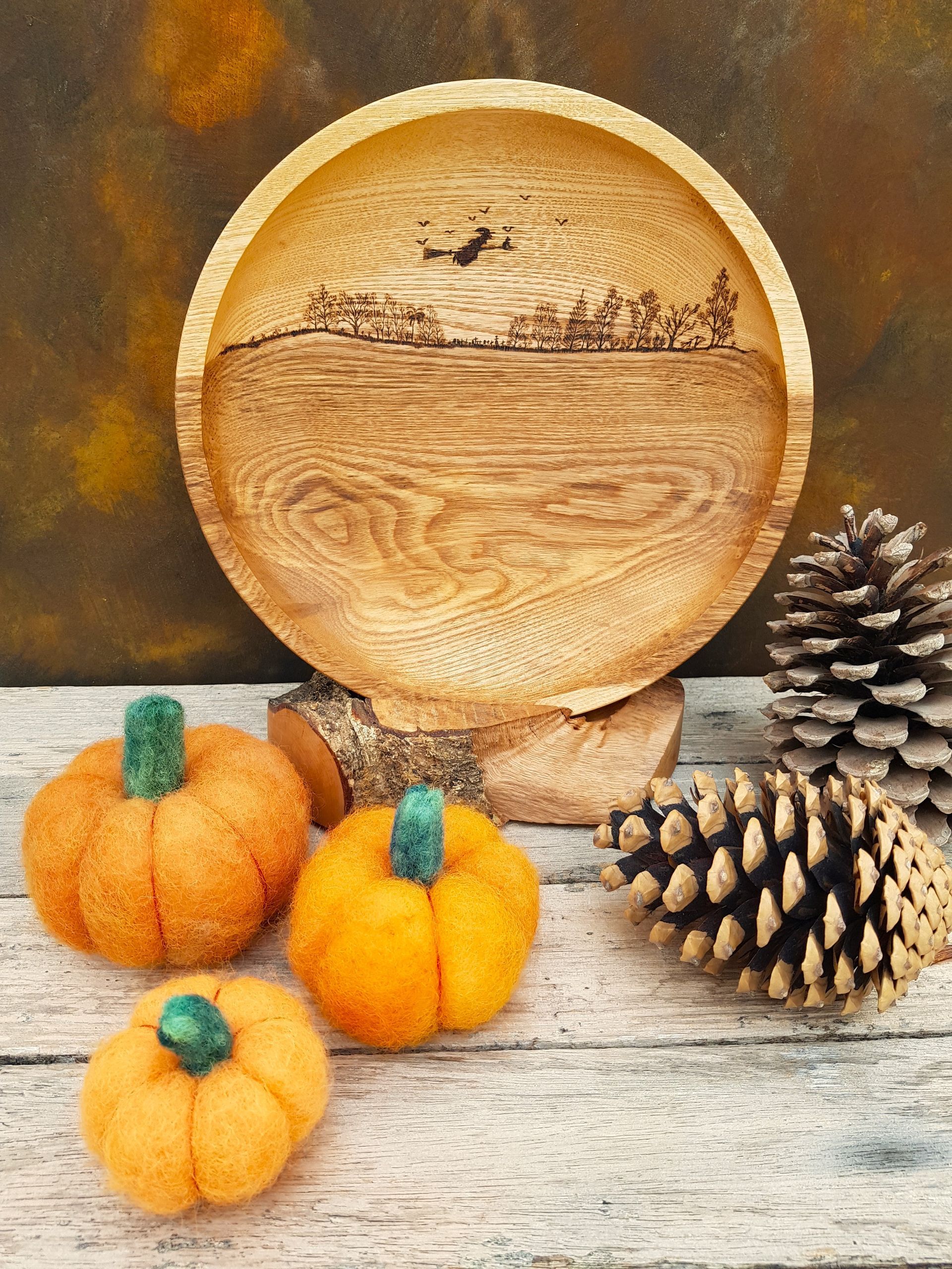 Autumn Inspired Large wooden bowl with striking grain pattern. Witch on broomstick flying above trees burnt onto the wood. 3 felted pumpkins and soe pinecones in foreground.
