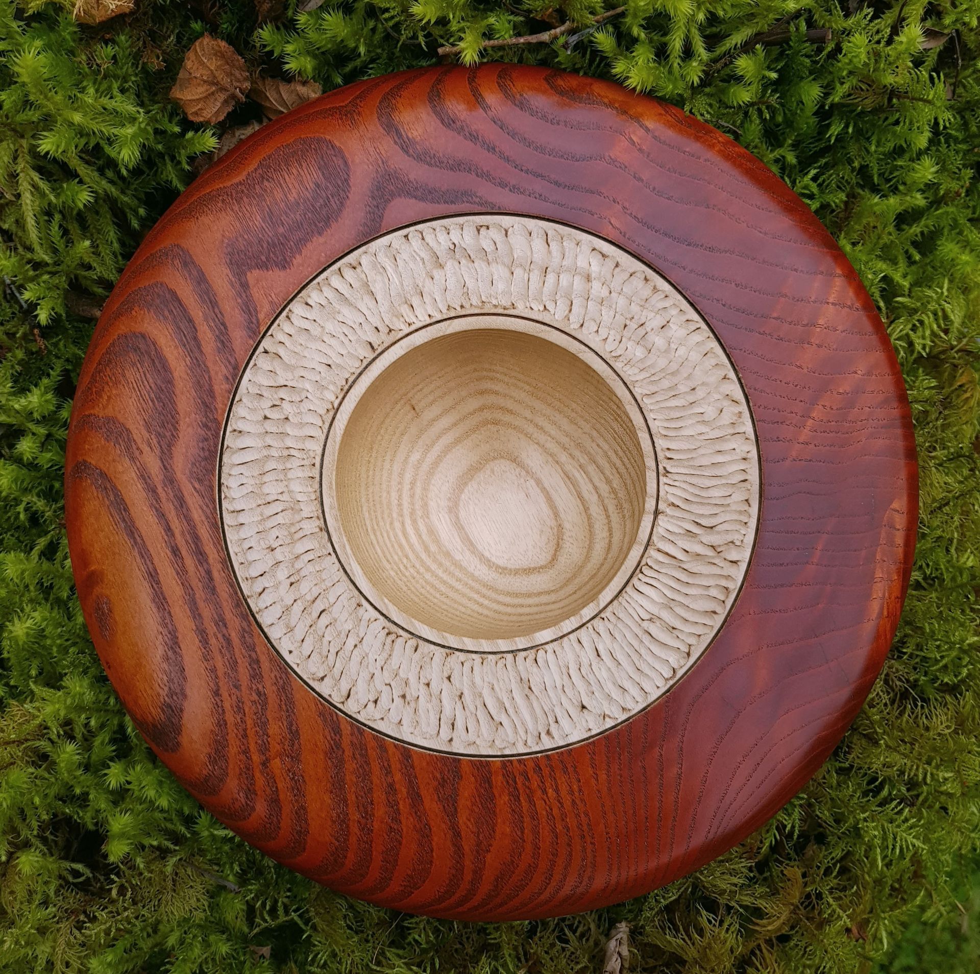 Ash bowl Large ash wood bowl, the outer section of the wide rim has been stained a burnt reddish brown and the scorched grain pattern is visible. The inner section of the rim is unstained but has been textured and the high spots have been scorched. The inside of the bowl is natural. Green moss background.