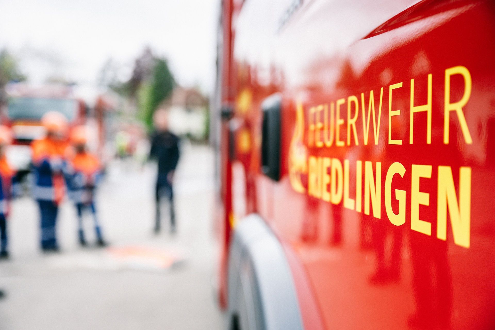 Nahaufnahme des Logos der Feuerwehr auf einem Feuerwehrfahrzeug. Im Hintergrund erkennt man mehrere Personen der Feuerwehr und Jugendfeuerwehr.
