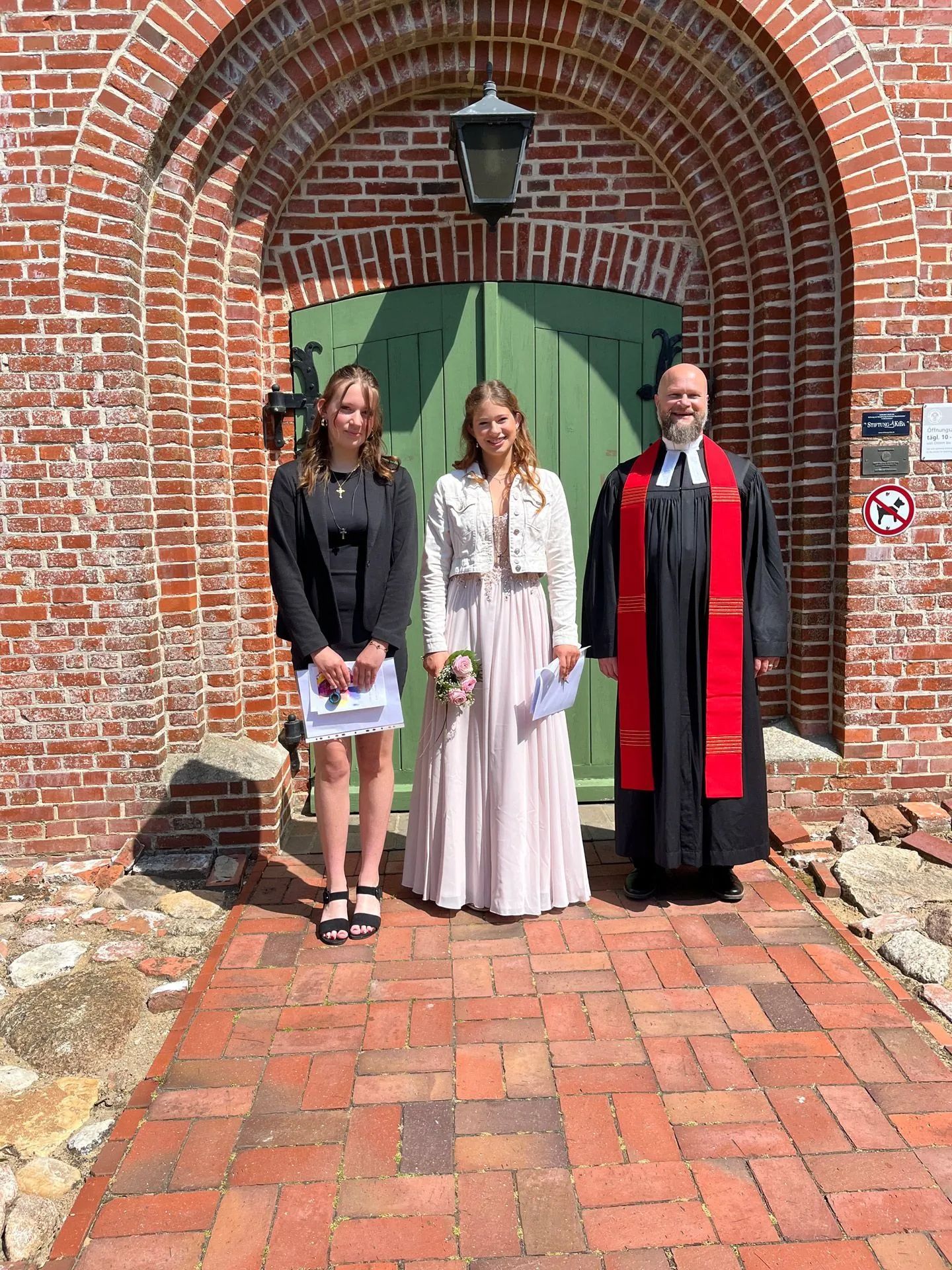 Konfirmation in der Kirche St. Martin in Vollerwiek, Eiderstedt. Konfirmation. Konfirmantinen mit Pastor vor Haupteingang.