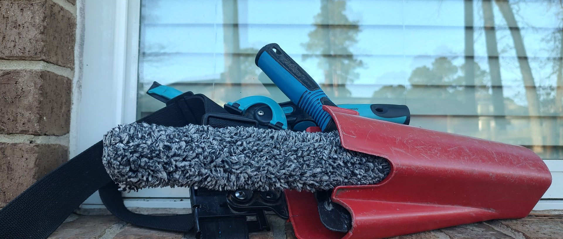 A professional window cleaning scrubber mop and soft-rubber squeegee sitting in a red bucket-on-a-belt (BOAB) on a residential window sill during a hand-washing service.