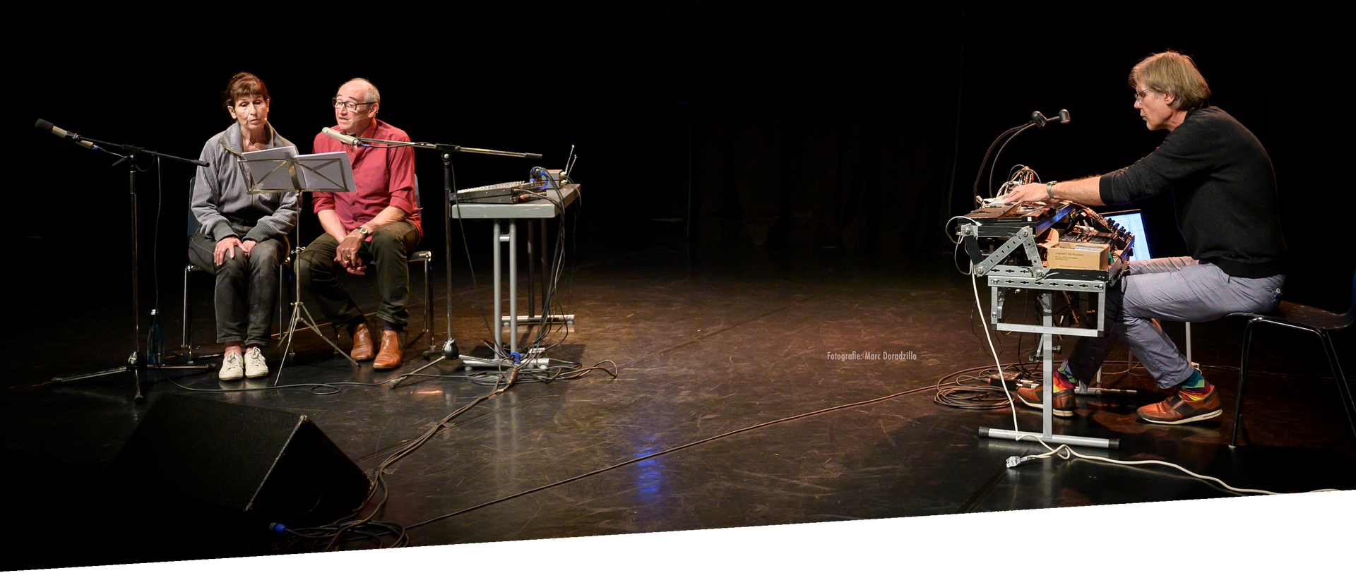 Zwei Personen lesen vor begleitet von einem Mann am Synthesizer