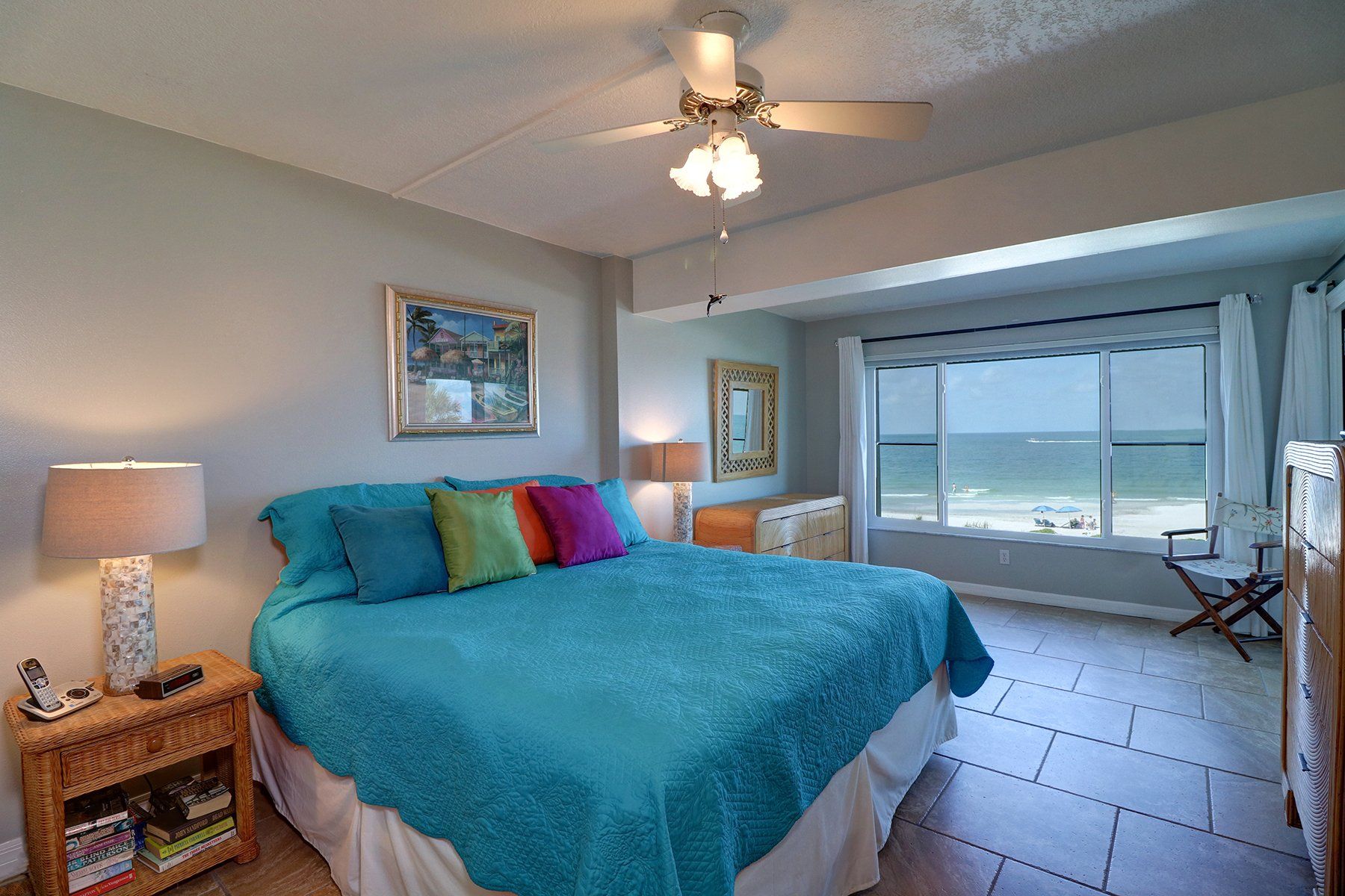 King bed with blue cover, multicolor pillows, bureau, mirror, lamp, chair and view of Gulf of Mexico
