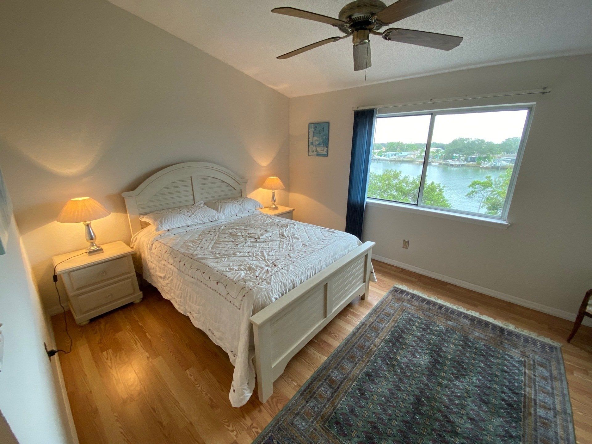 Queen bed with white wood frame, white bed cover, side tables and 2 lamps, blue window shade and Intracoastal view