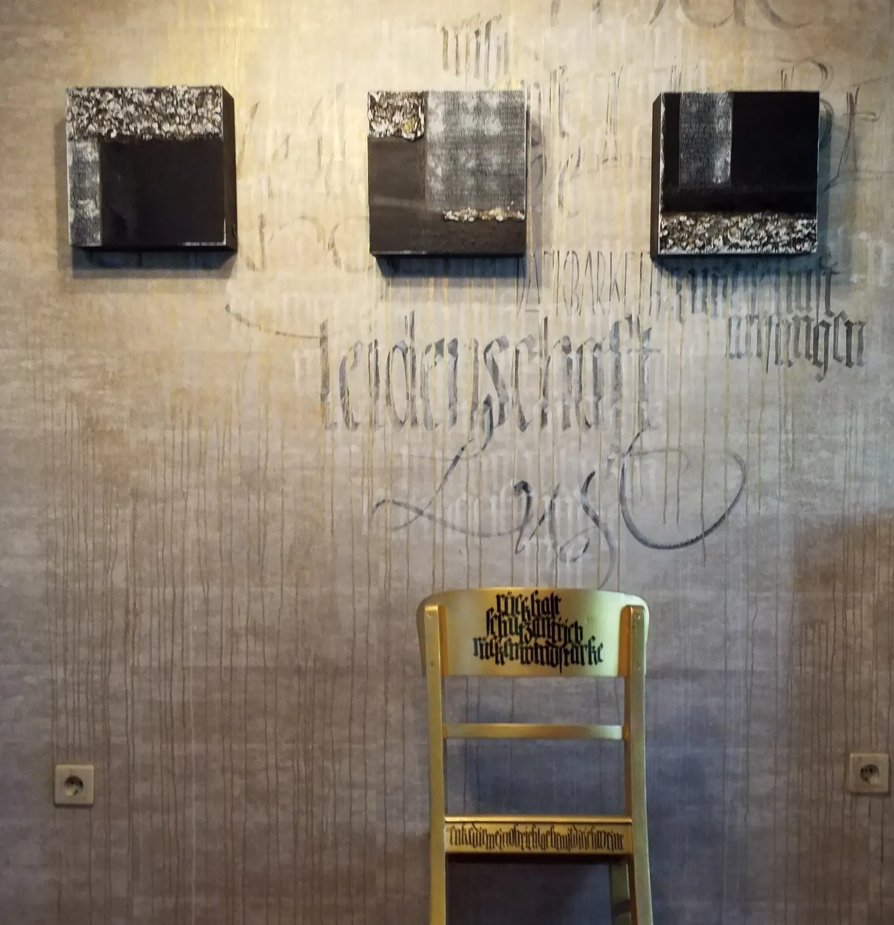 schriftkraft Ute Gräber, schriftkunz, beschriebener Stuhl, vor beschriebener Wand und 3er Serie Leinwände schwarz weiß