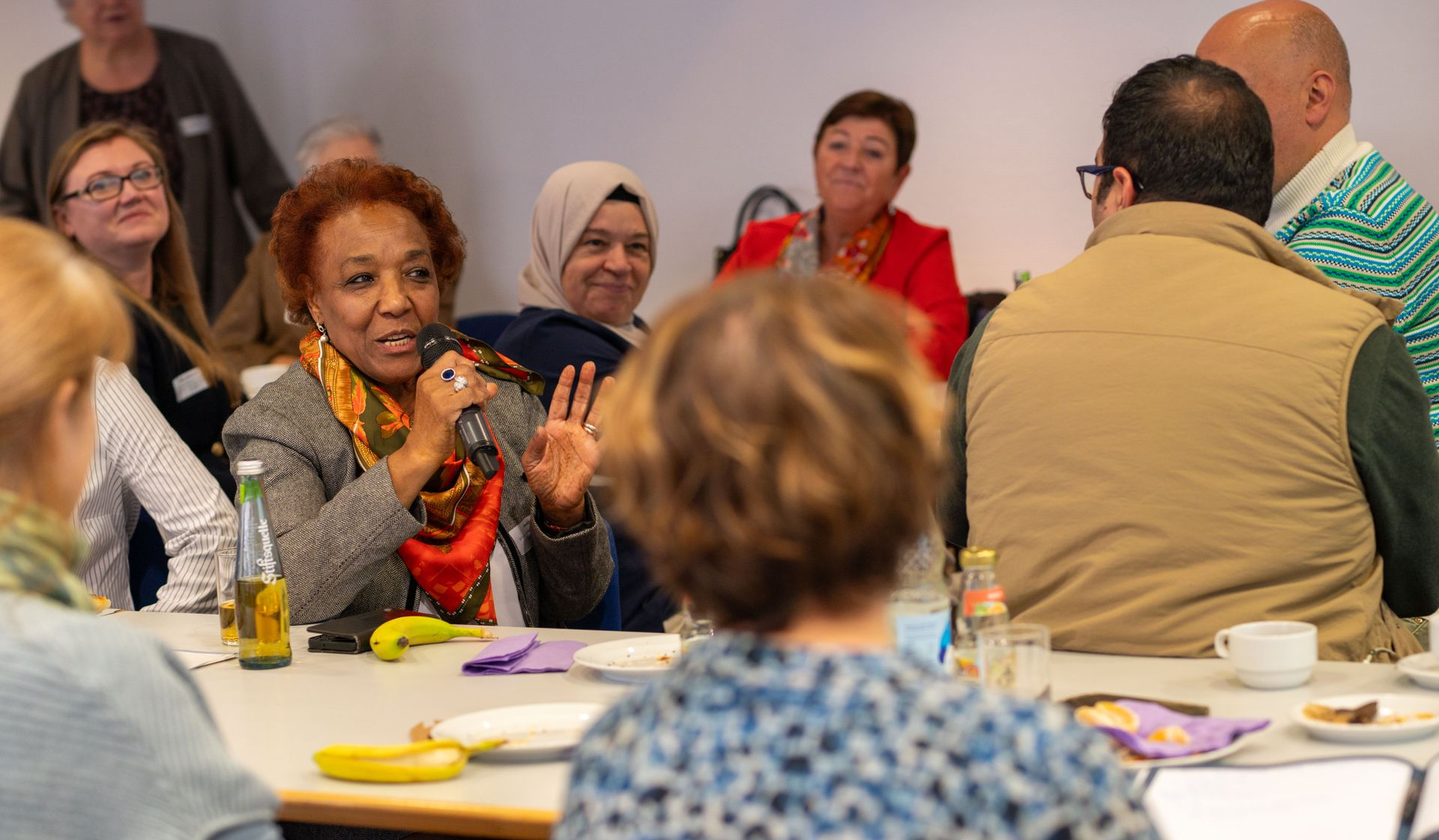 Frau spricht mit Mikrofon in Diskussionsrunde, Menschen sitzen an Tischen und hören aufmerksam zu.