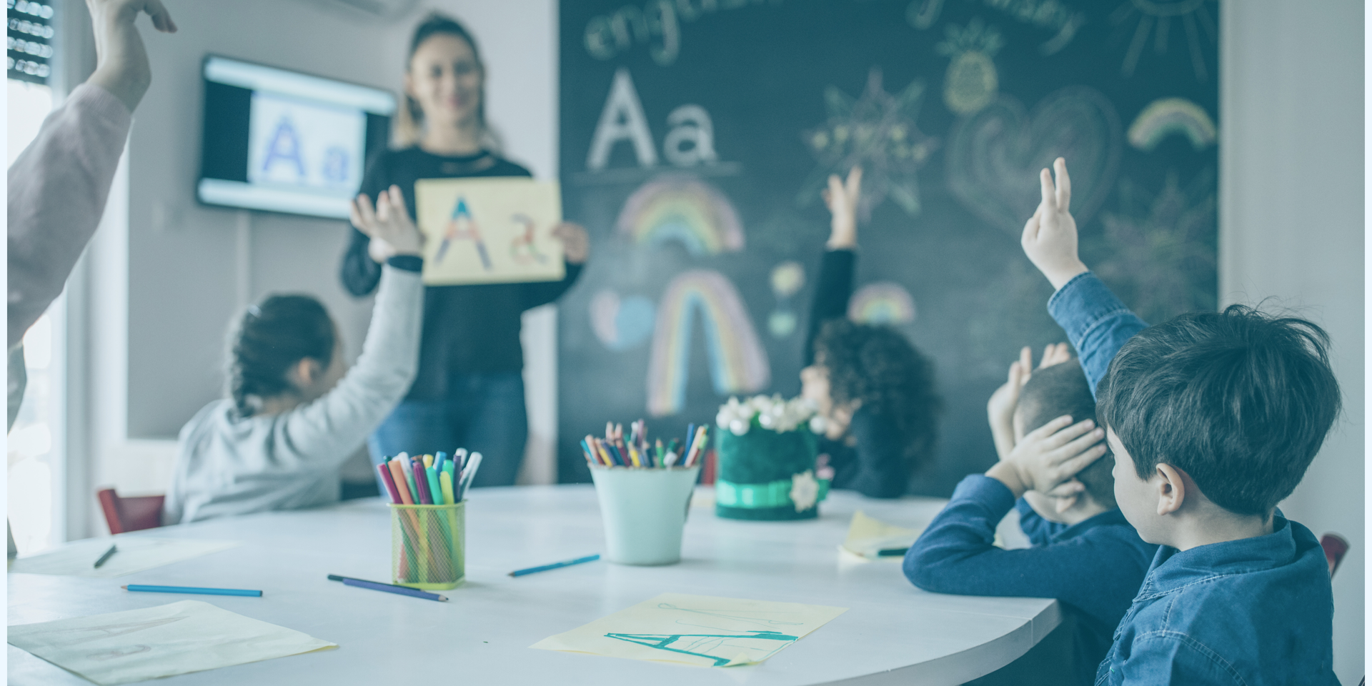 Kinder sitzen in einem Klassenzimmer und melden sich, während eine Lehrerin vorne Buchstaben zeigt.